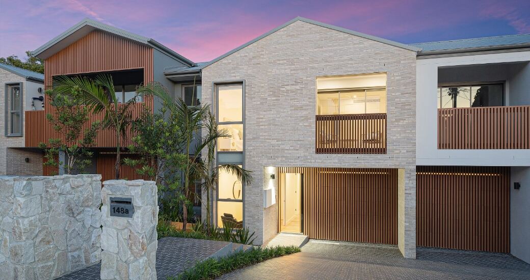 Dusk street view of the brick and timber-battened facade, showing the front entrance, garage door and stacked stone letterbox wall.
