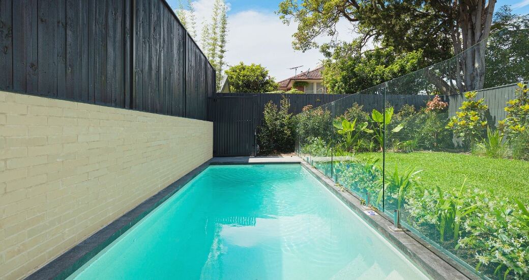 Long lap pool bordered by frameless glass fencing, dark timber fence and a sandstone-toned brick retaining wall.