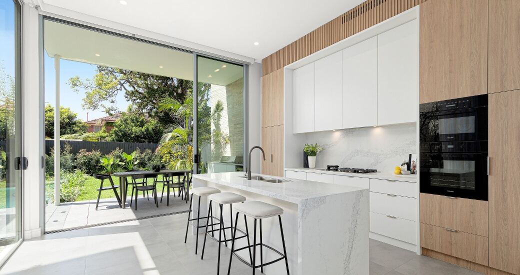 Kitchen with white stone island, black built-in oven tower and timber-panelled cabinetry, opening through sliding doors to the alfresco and garden.