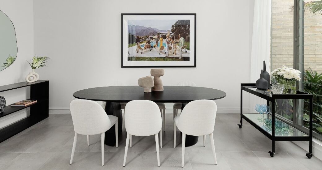 Dining room with a black oval table and boucle chairs beneath a framed photographic artwork, bar cart to the side.