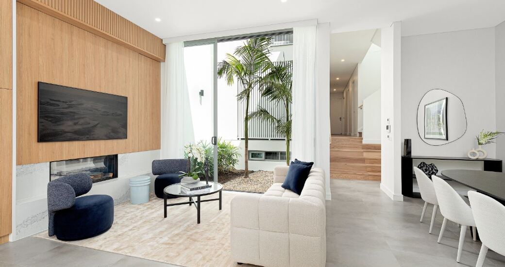 Open-plan living room with a timber feature wall, gas fireplace, wall-mounted TV and navy accent armchairs, opening to the dining area beyond.