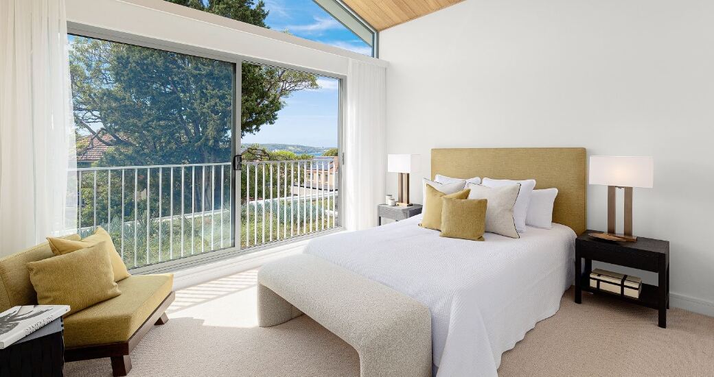 Primary bedroom with a vaulted timber-lined ceiling and floor-to-ceiling glazing opening onto a balcony with water views.