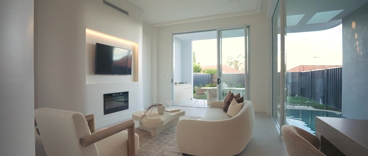 Living room with a wall-mounted fireplace and TV, curved glass room divider, and sliding doors opening onto the pool terrace