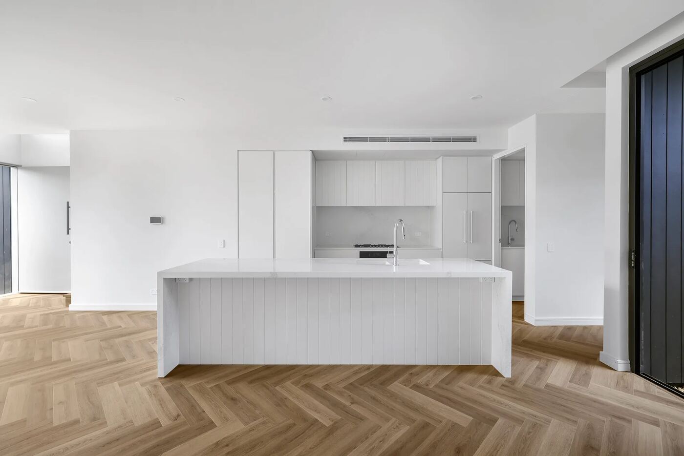 Minimalist white kitchen with waterfall-edge island bench, panelled cabinetry and integrated appliances above herringbone oak flooring