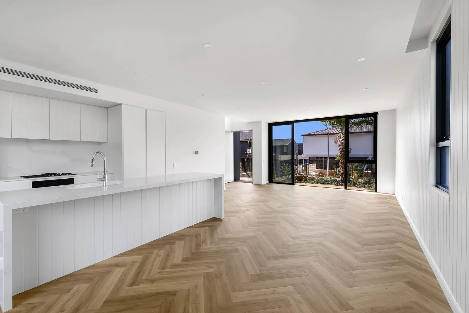Open-plan kitchen and living area with herringbone timber flooring opening onto a landscaped courtyard through full-height black-framed sliding doors